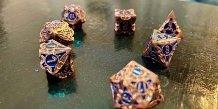Ett set med metalliska rollspels-tärningar med blå siffror ligger utspridda på ett blankt bord.