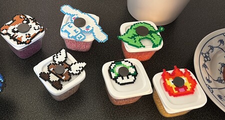 Sex små plastburkar med pärlplattor i form av spelkaraktärer på locken, står på ett svart bord.