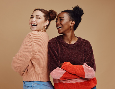 Två unga kvinnor står rygg mot rygg och skrattar mot kameran, mot beige bakgrund.