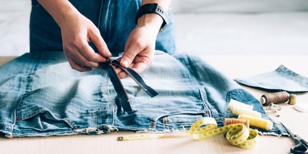 Person arbetar med att sy om ett par jeans, med dragkedja, måttband och sytråd på bordet.