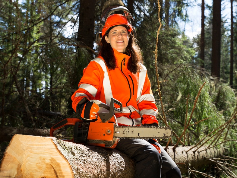 En kvinna i orange skyddskläder sitter på en fälld trädstam i skogen med en motorsåg i knät.