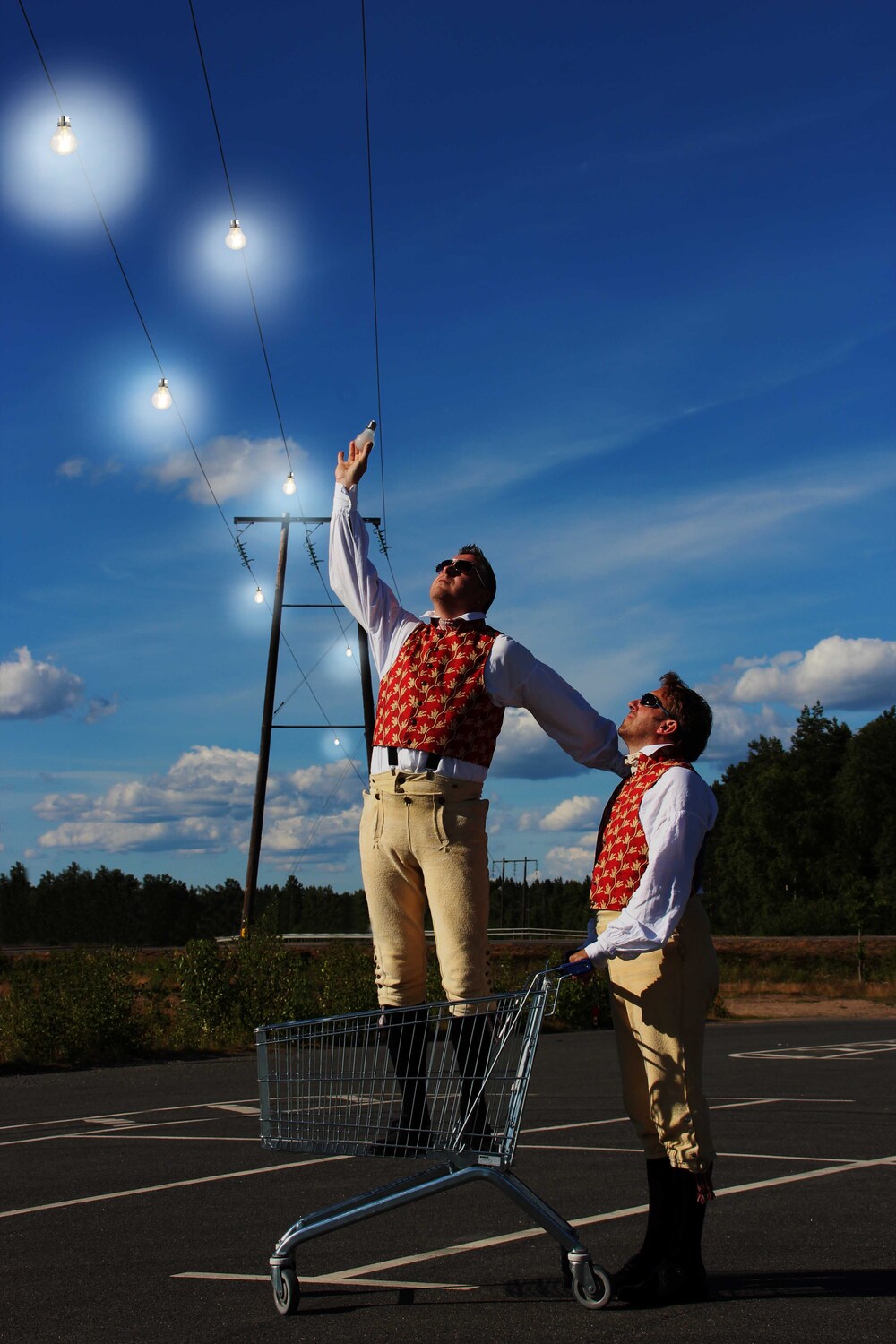 Två personer i färgglada västar står i en kundvagn på en tom parkering och sträcker sig mot glödlampor i himlen.