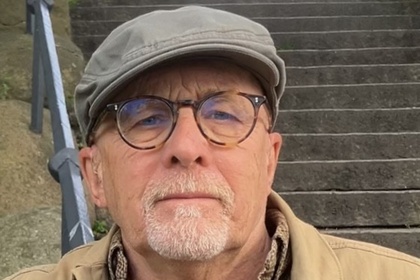 En person sitter utomhus vid en trappa, klädd i beige jacka och rutigt skjort, med keps och glasögon i bröstfickan.