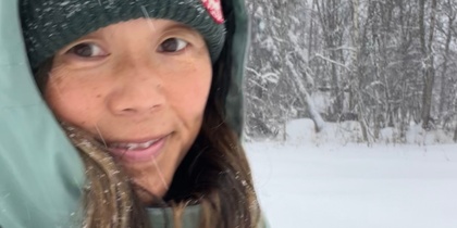 Person i vinterkläder står utomhus i ett snöigt landskap med träd i bakgrunden.