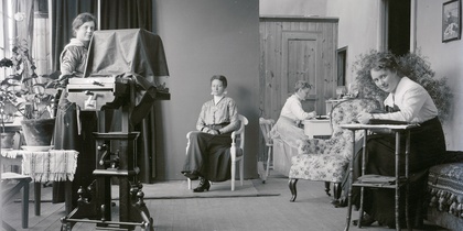Fyra kvinnor i ett rum, en poserar för ett gammaldags fotografi, andra arbetar och skriver.