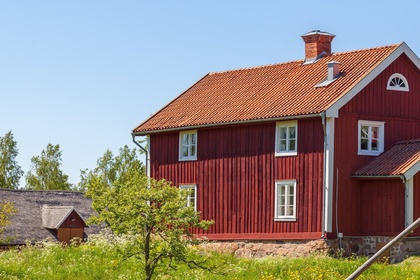 Två röda trähus med vita knutar på landet, omgivna av gräs, staket och blå himmel.