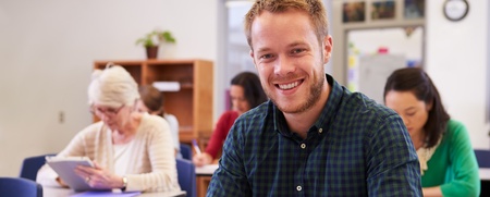 Leende man sitter vid skolbänk i klassrum, skriver i anteckningsbok. Andra vuxna studenter i bakgrunden.