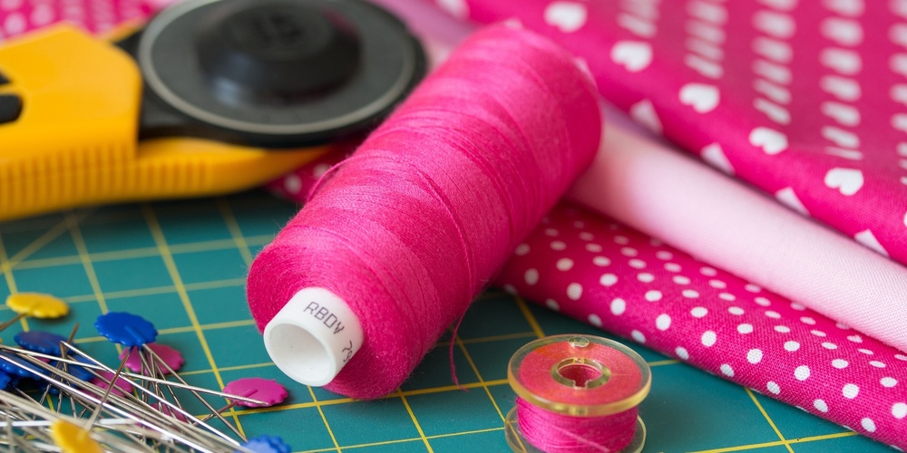 Närbild av sybehör: rosa tråd, knappnålar, rullkniv och rosa tyg med vita prickar och hjärtan.