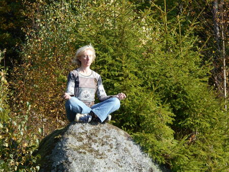 Person sitter i meditationsställning på en stor sten i skogen omgiven av gröna träd.