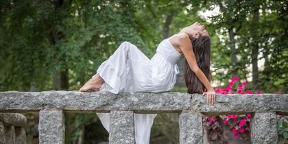Kvinna i vit klänning poserar liggande på ett stenräcke i en grönskande park.