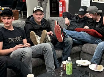 En grupp unga vuxna sitter i en soffa runt ett svart soffbord med drycker och tallrikar i ett vardagsrumsliknande rum.