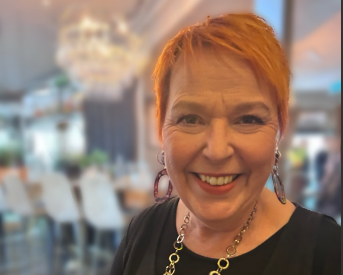 Person med kort, rött hår och svart klädsel står i en elegant restaurangmiljö med ljuskrona och vita stolar.