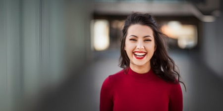 Leende person med långt mörkt hår och röd tröja står utomhus med suddig bakgrund.