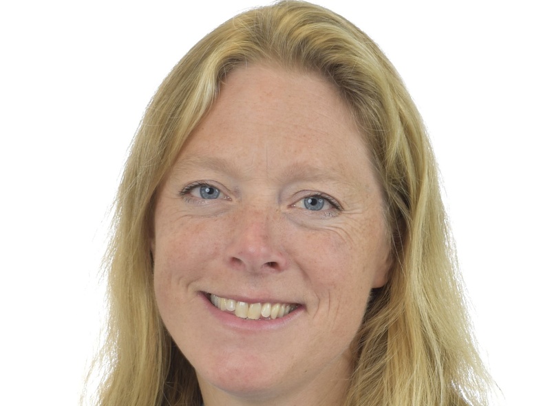 Kvinna med blont hår och ljusblå blus ler mot kameran mot vit bakgrund.