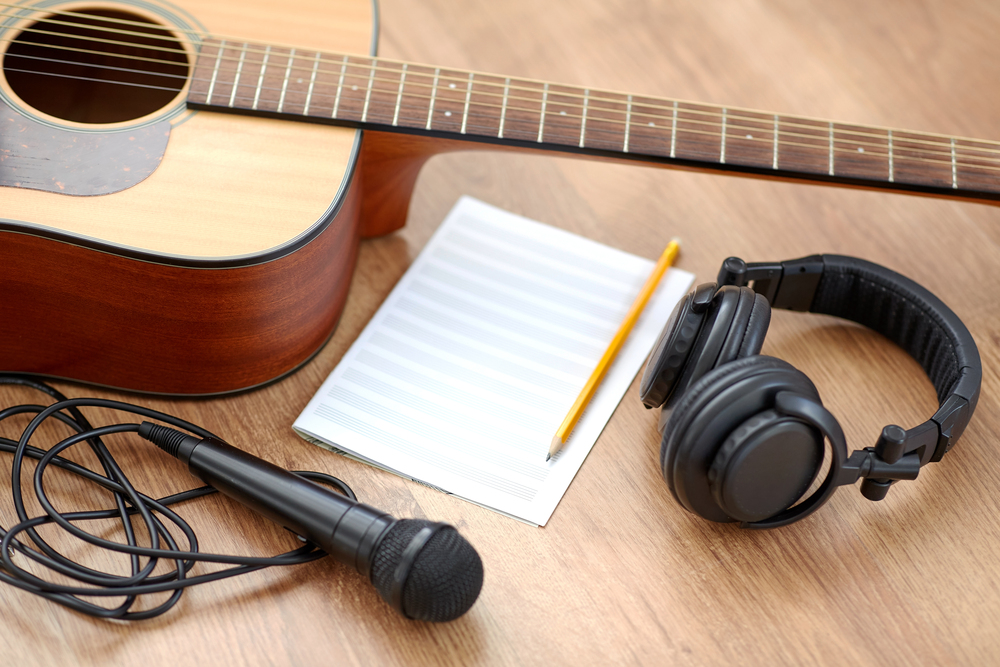 Akustisk gitarr, mikrofon, hörlurar, notpapper och penna på ett trägolv, redo för låtskrivning.