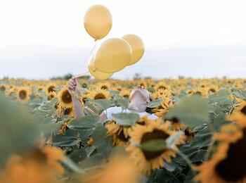 Person håller tre gula ballonger i ett fält av solrosor under en ljus himmel.
