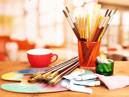 Ett bord med konstnärsmaterial som penslar, färgtuber, palett, en röd kopp och sudd i ett ljust rum.