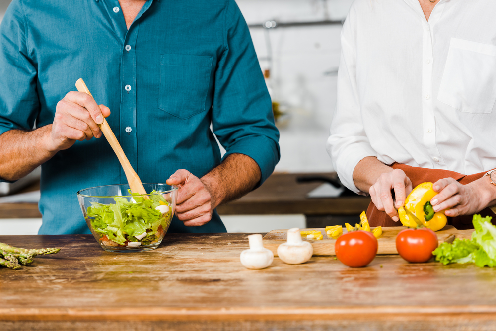 Två personer förbereder en sallad tillsammans i ett kök, en hackar grönsaker och en rör om i en skål.