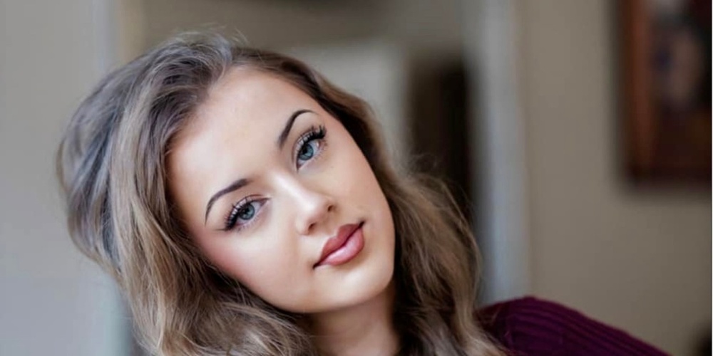 Ung kvinna med långt lockigt hår sitter avslappnat inomhus och tittar mot kameran.