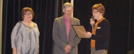 Tre vuxna personer står på en scen, en kvinna håller en mikrofon och ett diplom, de andra lyssnar.