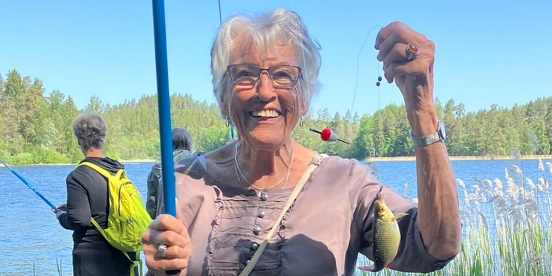 Äldre kvinna står på en brygga vid en sjö och håller upp en fisk hon fångat, med en annan person i bakgrunden.