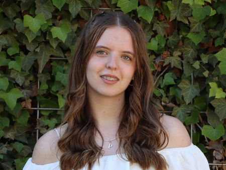 Ung kvinna med långt brunt hår och vit topp står framför en grön vägg av murgröna.