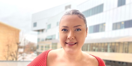 Kvinna i röd tröja står på en balkong med moderna byggnader i bakgrunden.