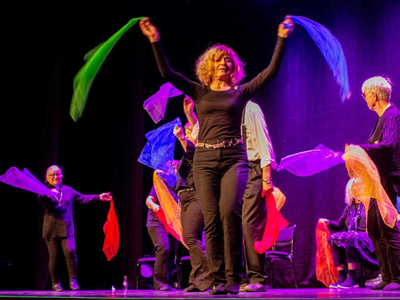 En grupp människor på scen viftar med färgglada tygsjalar under en föreställning med lila scenbelysning.