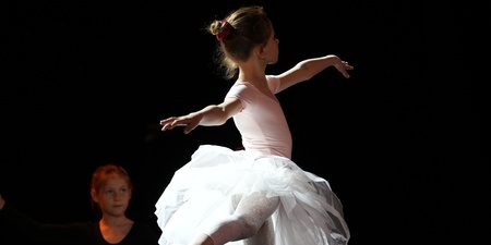 En ung ballerina i vit tyllkjol dansar på scenen med armarna utsträckta, belyst av scenljus.