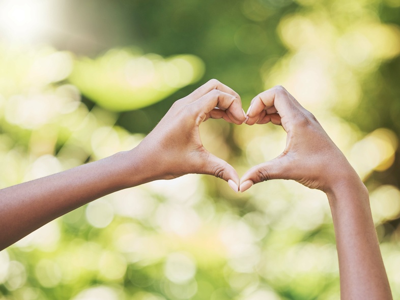 Två händer formar ett hjärta mot en suddig, grön bakgrund utomhus.