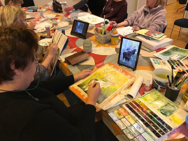 Flera personer sitter runt ett bord och målar akvareller med hjälp av surfplattor som visar djurbilder.