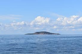 En liten ö syns i fjärran omgiven av ett lugnt blått hav under en klar himmel med moln.