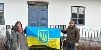 Två personer står utanför en byggnad och håller upp en ukrainsk flagga med vapensköld.