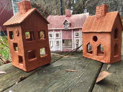 Tre små modellhus i lera och trä står på en väderbiten träyta utomhus med gräs och träd i bakgrunden.