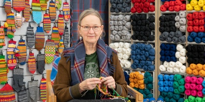 En kvinna sitter och stickar i en garnbutik omgiven av färgglada garnnystan och stickade vantar på väggen.