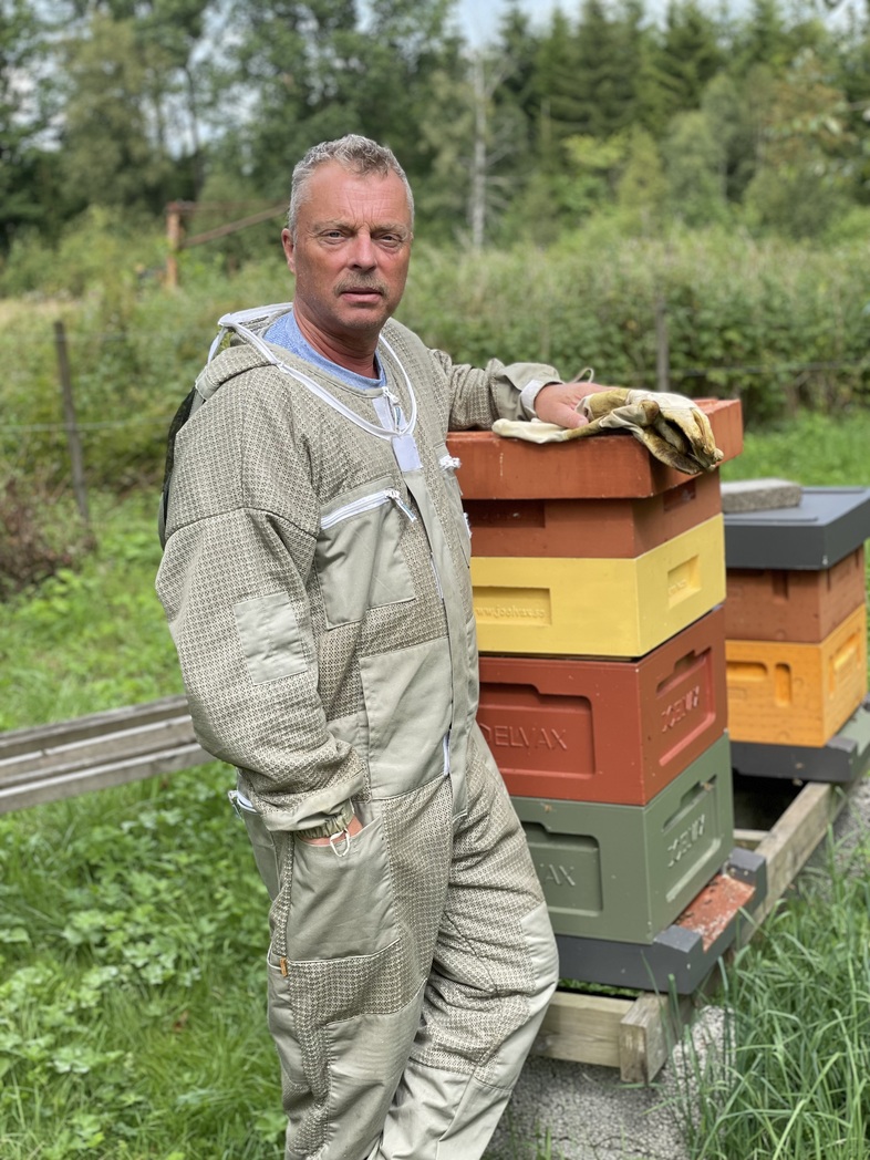 En man i biodlardräkt står bredvid färgglada bikupor utomhus på en grön äng.