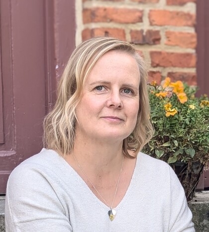 Person med blont hår sitter utomhus framför en tegelvägg och blommor, ansiktet är suddat.