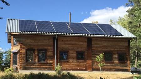 Trähus med solpaneler på taket, omgiven av träd och blå himmel.