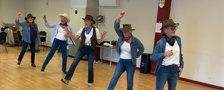 Fem personer dansar linedance inomhus, klädda i cowboyhattar, jeans och scarfar.