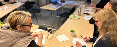 Flera personer sitter vid bord i ett mötesrum och deltar i ett hybridmöte med en person på storbildsskärm.