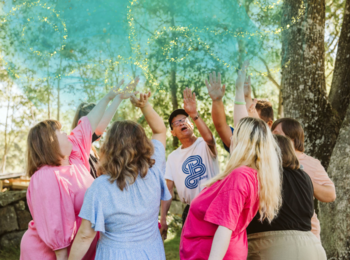 En grupp människor står i en ring utomhus och kastar färgat pulver i luften under träd.