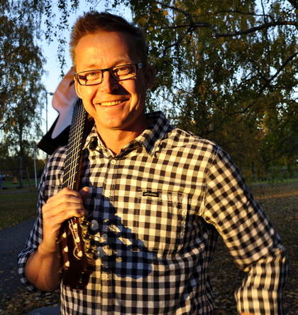Leende man med glasögon och gitarr på axeln står utomhus i kvällssol, klädd i rutig skjorta.