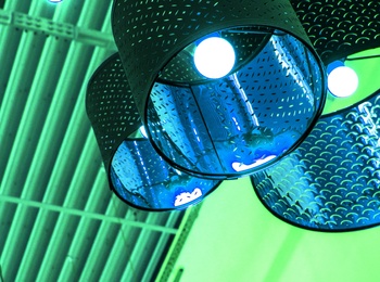 Flera taklampor med industriell design hänger från ett tak med synliga balkar, belysta i grönt och blått ljus.