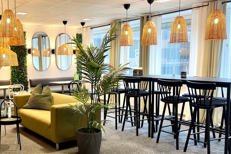 Modern loungeyta med gröna växter, soffor, höga bord och stolar samt stora fönster med naturligt ljus.