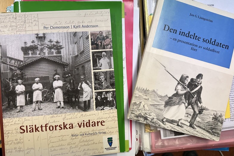 Två böcker på svenska om släktforskning och soldatliv ligger på ett bord bland papper och mappar.