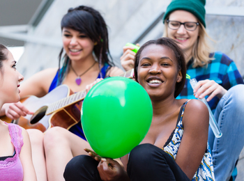 Fyra unga personer sitter tillsammans utomhus, en spelar gitarr och en annan håller en grön ballong.