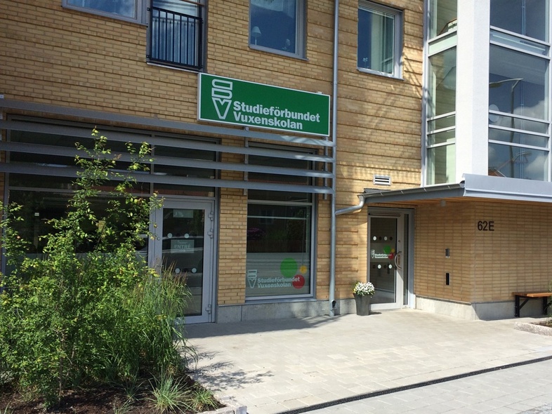 Entré till en modern byggnad med skylt för Studieförbundet Vuxenskolan, omgiven av växter och gångväg.