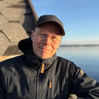 Person med mörk jacka och keps sitter utomhus vid vatten, med ett hus i bakgrunden under klarblå himmel.