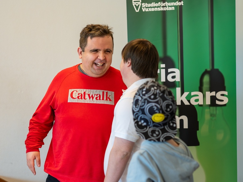 Tre personer samtalar framför en grön skylt med texten 'Studieförbundet Vuxenskolan'. En person bär röd tröja med texten 'Catwalk'.