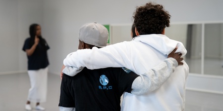 Två personer står med armarna om varandra, sedda bakifrån, i ett ljust rum. En tredje person syns i bakgrunden.